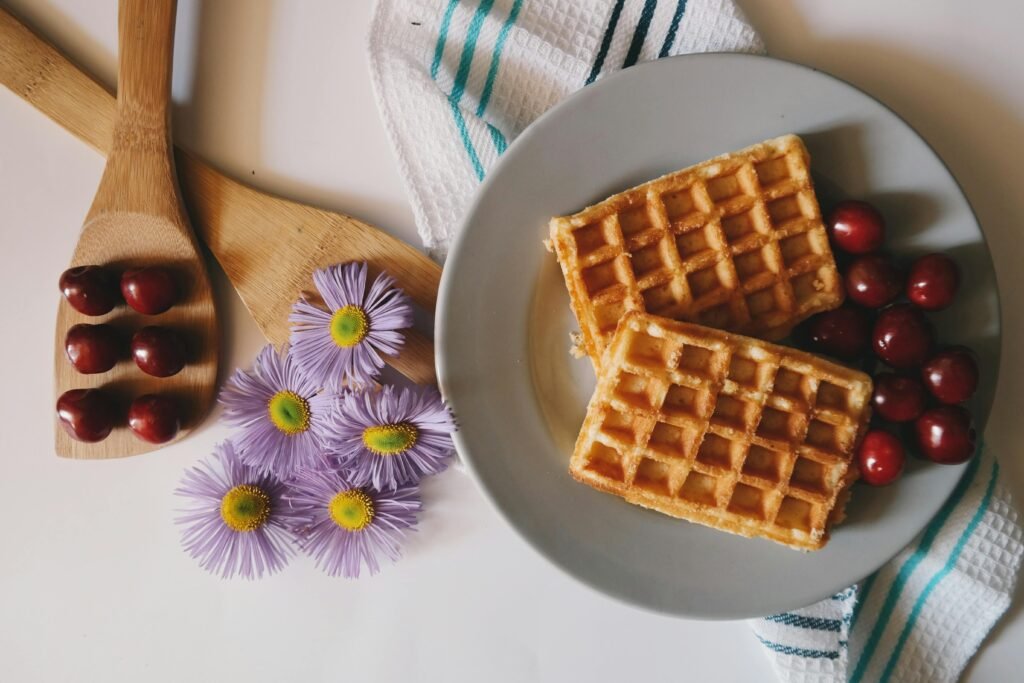 Top view of waffles with cherries and daisies on a plate, styled with a wooden spoon and towel.